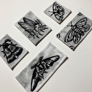 Pode incluir: Cinco patches de tecido com ilustrações de mariposas pretas sobre um fundo cinza. As mariposas são retratadas em várias poses, com padrões de asas detalhados. Cada patch é retangular ou quadrado, com uma borda costurada.