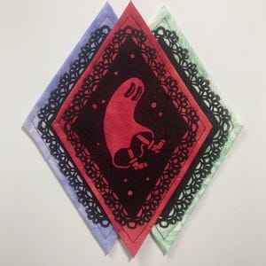 May include: Three diamond-shaped fabric patches layered together. The left is light purple, the centre is red, and the right is light green. The centre red patch features a black lace-like border and a central graphic of a red ghost figure.
