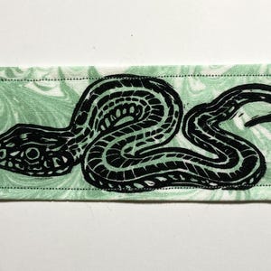 May include: A rectangular fabric patch featuring a black ink block print of a coiled snake with a forked tongue. The background fabric is a light mint green with a white marbled pattern, and the entire piece is finished with a black stitched border.