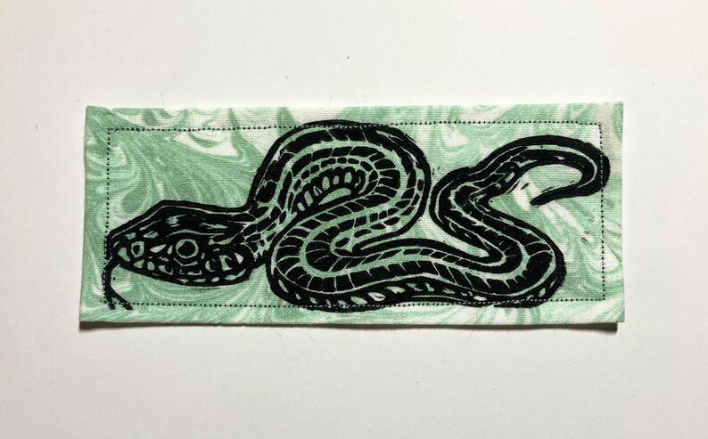 May include: A rectangular fabric patch featuring a black ink block print of a coiled snake with a forked tongue. The background fabric is a light mint green with a white marbled pattern, and the entire piece is finished with a black stitched border.