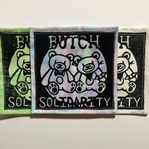 May include: Three square fabric patches featuring a black ink block print of two teddy bears and the text BUTCH SOLIDARITY. The centre patch is on a pastel tie-dye fabric, the left is on light green, and the right is on an off-white fabric.