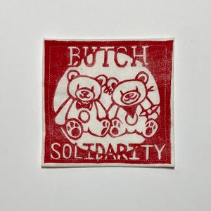 May include: A square patch in red and white featuring the words "BUTCH SOLIDARITY" and two cartoon teddy bears. The bears are white with black outlines and details, and the text is in a bold, sans-serif font.