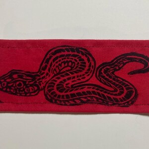 May include: A rectangular red fabric patch featuring a black ink-style print of a coiled snake. The design shows the snake in a side profile with detailed scale patterns across its body. The patch has visible stitched borders along all four edges.