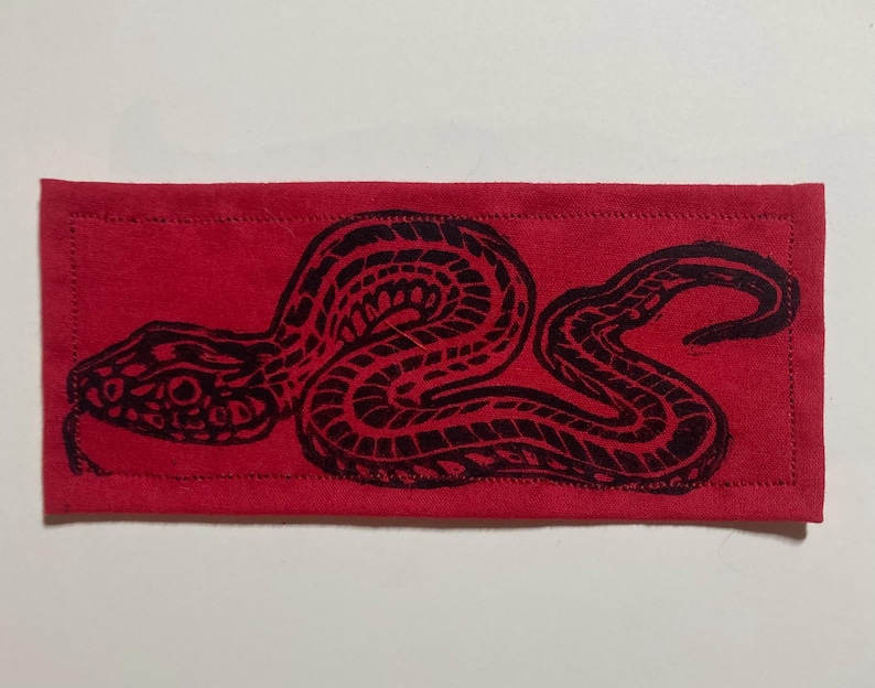 May include: A rectangular red fabric patch featuring a black ink-style print of a coiled snake. The design shows the snake in a side profile with detailed scale patterns across its body. The patch has visible stitched borders along all four edges.