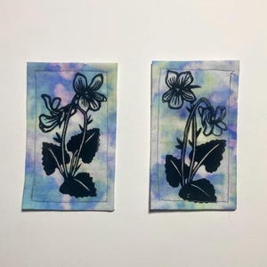 Könnte beinhalten: Zwei rechteckige Stoffflicken mit einem Batik-Effekt in Blau, Lila und Grün. Jeder Flicken zeigt ein schwarzes Blumendesign mit einer Blume und Blättern. Die Ränder sind mit hellem Faden genäht.