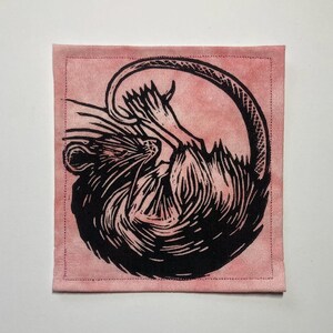 K&ouml;nnte beinhalten: Ein quadratischer Stoffaufn&auml;her mit einem schwarzen Linoldruck einer zusammengerollten Ratte auf einem altrosa Hintergrund. Die Ratte ist in einem stilisierten, illustrativen Stil mit sichtbarer Fellstruktur und einem langen Schwanz dargestellt.
