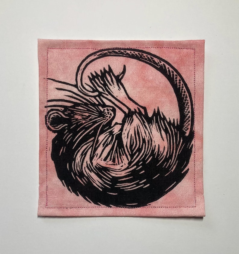 K&ouml;nnte beinhalten: Ein quadratischer Stoffaufn&auml;her mit einem schwarzen Linoldruck einer zusammengerollten Ratte auf einem altrosa Hintergrund. Die Ratte ist in einem stilisierten, illustrativen Stil mit sichtbarer Fellstruktur und einem langen Schwanz dargestellt.