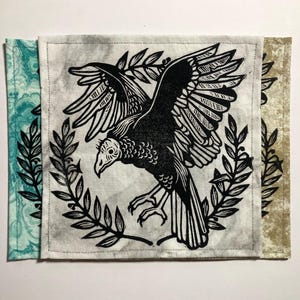 May include: Square fabric art with a black and white vulture illustration, wings spread, within a laurel wreath. The central image is on a light grey background, with teal and tan patterned fabric edges.