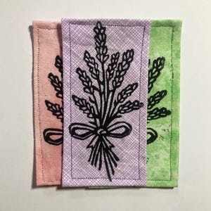 Peut inclure: Trois dessous de verre rectangulaires en tissu rose, lavande et vert. Chaque dessous de verre présente une illustration noire d'un bouquet de lavande noué avec un nœud. Les dessous de verre sont probablement faits à la main et ont une bordure cousue.