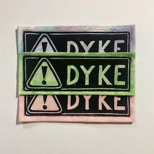 Puede incluir: Tres parches rectangulares de tela apilados verticalmente. Cada parche presenta un fondo negro con un triángulo blanco que contiene un signo de exclamación y la palabra DYKE en letras mayúsculas blancas. Los bordes son de colores pastel.
