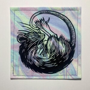 K&ouml;nnte beinhalten: Ein quadratischer Stoffaufn&auml;her mit einem schwarzen Blockdruck eines zusammengerollten Rattenmotivs. Der Hintergrundstoff zeigt ein pastellfarbenes Batikmuster in Rosa, Blau und Gelb. Der Aufn&auml;her hat einen gen&auml;hten Rand an allen vier Seiten.
