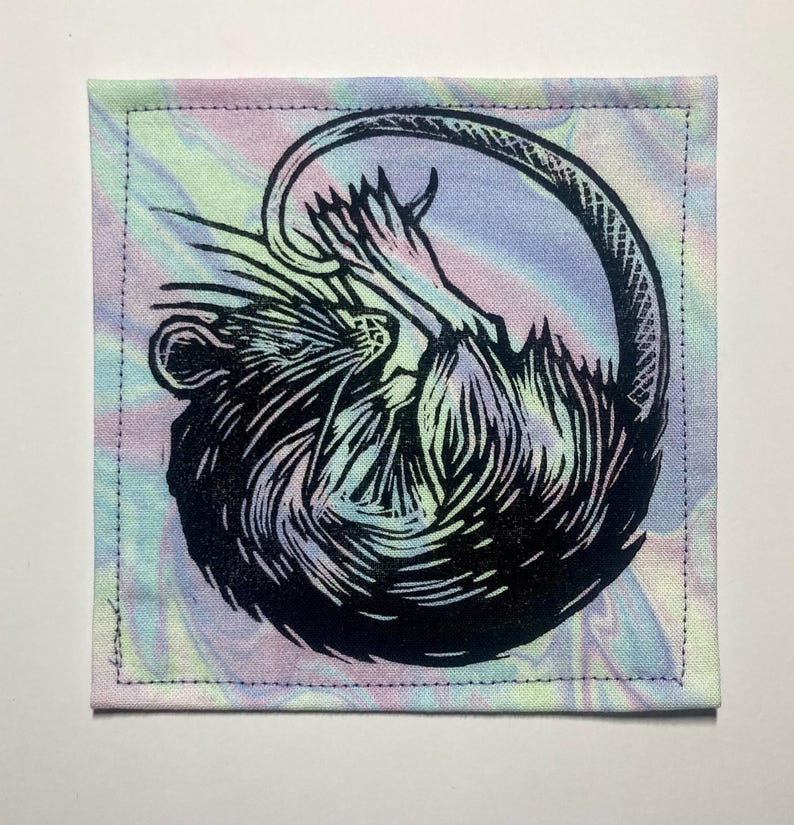 K&ouml;nnte beinhalten: Ein quadratischer Stoffaufn&auml;her mit einem schwarzen Blockdruck eines zusammengerollten Rattenmotivs. Der Hintergrundstoff zeigt ein pastellfarbenes Batikmuster in Rosa, Blau und Gelb. Der Aufn&auml;her hat einen gen&auml;hten Rand an allen vier Seiten.