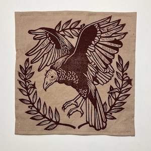 Könnte beinhalten: Quadratische, beigefarbene Stoffserviette mit einer dunkelbraunen Illustration eines Geiers mit ausgebreiteten Flügeln, umrahmt von einem Lorbeerkranz. Die Serviette hat einen genähten Rand.