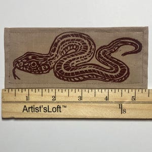 May include: A rectangular tan fabric patch featuring a dark brown block-printed snake design. The patch is positioned above a wooden ruler, showing a total length of approximately 6 inches. The edges of the fabric are stitched with a straight border.
