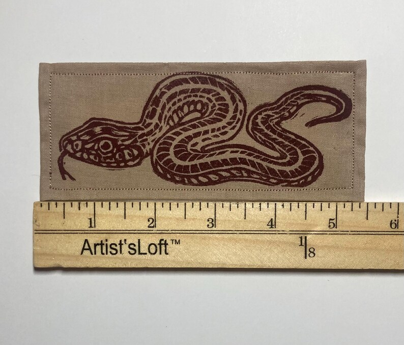 May include: A rectangular tan fabric patch featuring a dark brown block-printed snake design. The patch is positioned above a wooden ruler, showing a total length of approximately 6 inches. The edges of the fabric are stitched with a straight border.