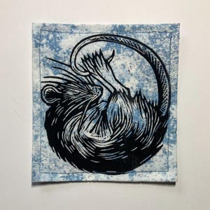 K&ouml;nnte beinhalten: Ein quadratischer Stoffaufn&auml;her mit einem schwarzen Linoldruck einer zusammengerollten Ratte auf hellblau-wei&szlig; gesprenkeltem Hintergrund. Das Motiv ist von einer einfachen Naht entlang der Kanten des quadratischen Textilst&uuml;cks umrandet.