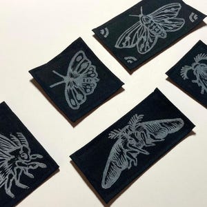May include: A collection of six black fabric patches, each featuring a detailed silver moth or butterfly design. The patches are rectangular and arranged on a white surface, showcasing the insect illustrations.