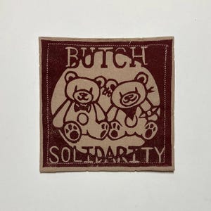 May include: A square patch with a maroon border and a beige centre. The patch features two cartoon teddy bears holding hands, with the words "BUTCH SOLIDARITY" in maroon text above and below the bears.