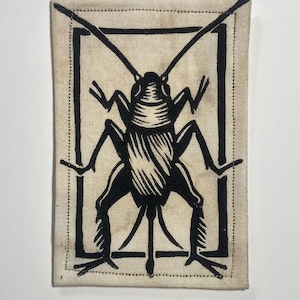 Peut inclure: Un écusson rectangulaire avec un dessin d'insecte noir à l'intérieur d'un cadre rectangulaire noir. L'insecte a des pattes, des antennes et des segments corporels détaillés. L'écusson est sur un tissu beige avec une bordure pointillée.