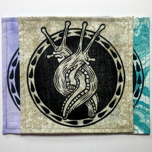 May include: Three square fabric coasters featuring a black ink block print of two intertwined slugs inside a circular border with teardrop shapes. The coasters have different background colours: a light purple, a mottled beige, and a teal marbled pattern.