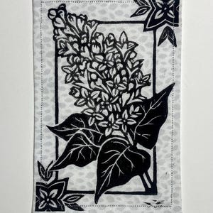 Op de afbeelding: Zwart-wit bloemen linoleumsnede print van een seringen tak met bladeren, omlijst door een zwarte rand. De achtergrond is een lichtgrijs en wit gestippeld patroon. De print staat op een rechthoekig stuk stof.