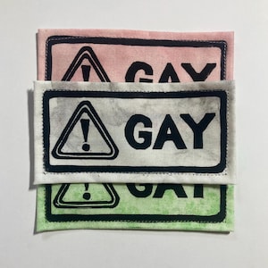 Peut inclure: Trois écussons rectangulaires en tissu empilés verticalement. Chaque écusson présente une icône de panneau d'avertissement noire avec un point d'exclamation dans un triangle, suivie du mot GAY en lettres capitales noires grasses. Les tissus sont rose, blanc et vert clair.