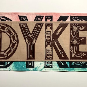 Può includere: Un'opera d'arte tessile a strati con la parola "DYKE" in lettere a blocchi marroni. La "D" presenta un design a catena, la "Y" ha una cravatta e la "E" accenti floreali. Lo sfondo include strati rosa, turchese e neri.