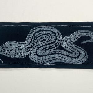 May include: A rectangular black fabric patch featuring a white screen-printed graphic of a coiled snake with intricate scale patterns. The patch has a visible white stitched border running along the perimeter of the rectangular shape.