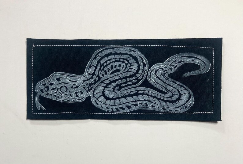 May include: A rectangular black fabric patch featuring a white screen-printed graphic of a coiled snake with intricate scale patterns. The patch has a visible white stitched border running along the perimeter of the rectangular shape.