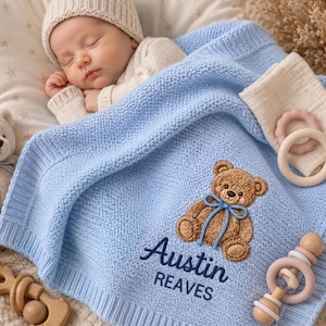 May include: A light blue knitted baby blanket featuring an embroidered brown teddy bear with a blue ribbon and the text Austin REAVES. The blanket is accompanied by a small teddy bear, a wooden rattle, and a silicone ring toy.