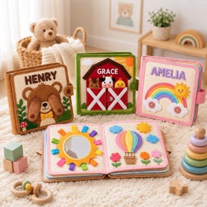 May include: Three square fabric activity books with personalized covers featuring a bear named Henry, a barn named Grace, and a rainbow named Amelia. One book is open, displaying a sun mirror and a hot air balloon with colorful felt flowers.