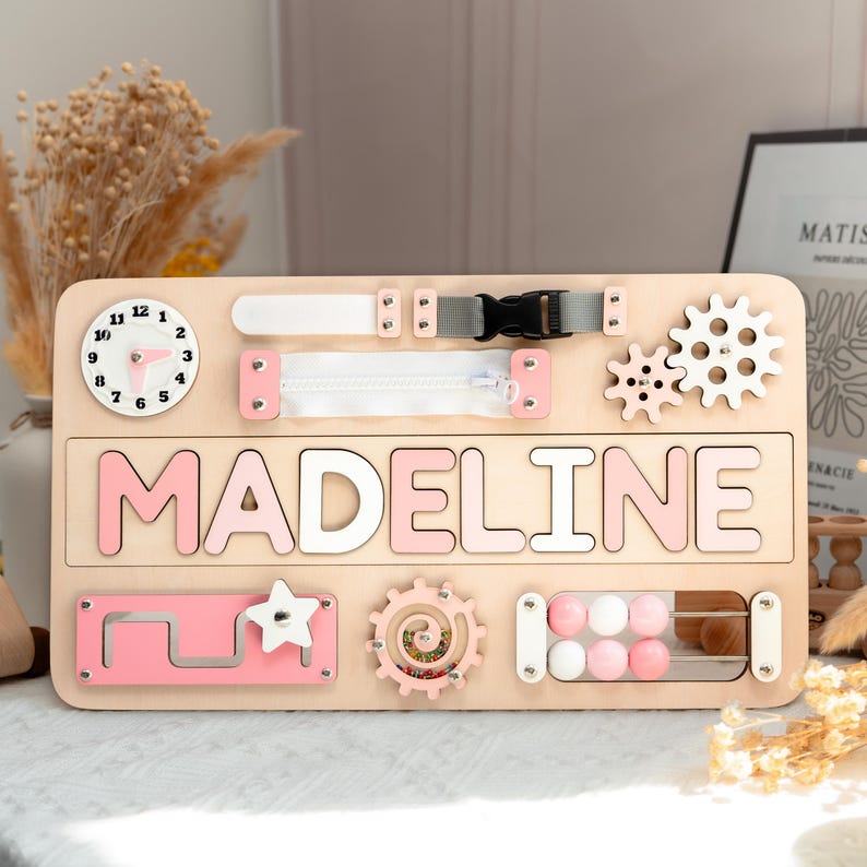 A wooden name board with the name "MADELINE" written on it, surrounded by various colorful and interactive elements such as gears, wheels, and a clock.
