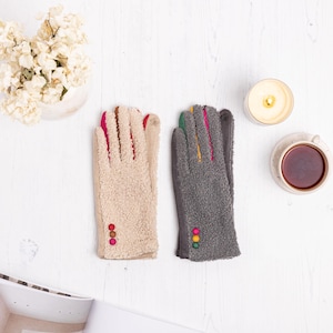 May include: Two pairs of gloves. One is beige with pink and red details, the other is grey with green, yellow, and pink accents. Each glove has three decorative buttons. The gloves are on a white surface.