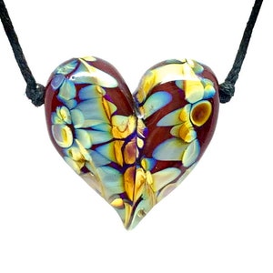 Può includere: Un ciondolo di vetro a forma di cuore con un design astratto colorato in tonalità di blu, oro e rosso. Il ciondolo è infilato su un cordoncino nero.
