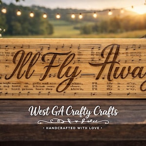 Peut inclure: Panneau en bois rectangulaire avec l'inscription gravée "I'll Fly Away" dans une police décorative, sur fond de notes de musique. Le panneau est en bois clair et porte le texte "West GA Crafty Crafts" en bas.