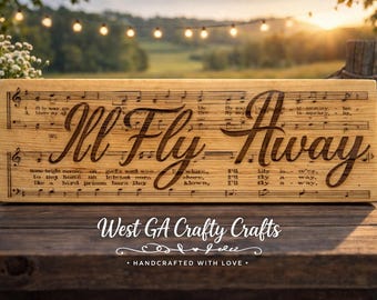 May include: Wooden sign with the engraved phrase "I'll Fly Away" in a decorative font, set against a backdrop of musical notes. The sign is made of light-colored wood and features the text "West GA Crafty Crafts" at the bottom.