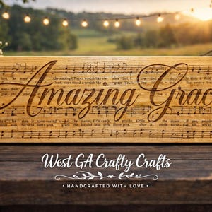 Op de afbeelding: Rechthoekig houten bord met de woorden "Amazing Grace" en muzieknoten gegraveerd. Het bord is gemaakt van lichtgekleurd hout. De tekst "West GA Crafty Crafts" staat onderaan.