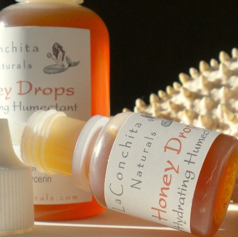May include: Two amber glass bottles with white labels. The smaller bottle is labeled "La Conchita Naturals Honey Drops Hydrating Humectant". The larger bottle is labeled "La Conchita Naturals Honey Drops Hydrating Humectant".