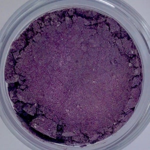 May include: A jar filled with a deep purple powder. The powder has a fine, granular texture and appears to be a cosmetic product, such as eyeshadow or pigment. The clear jar allows the color to be fully visible.