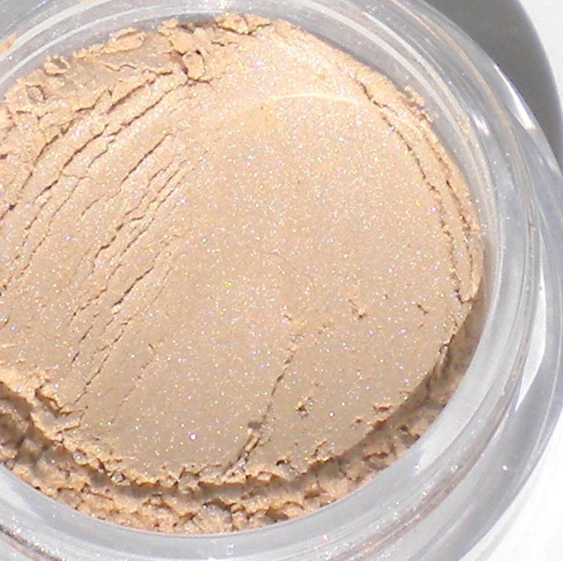 Op de afbeelding: Een close-up van een kleine, ronde container met losse, lichtbeige oogschaduw met een glinsterende afwerking.