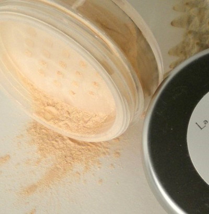 K&ouml;nnte beinhalten: Ein durchsichtiges Plastikglas mit losem, hellbeigem Puder mit einem Siebaufsatz, teilweise ge&ouml;ffnet, der auf eine wei&szlig;e Oberfl&auml;che versch&uuml;ttet wird. Ein silberner Deckel liegt neben dem Glas. Das Puder scheint ein Kosmetikprodukt zu sein.