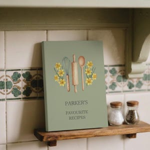 May include: A sage green recipe book with the text "PARKER'S FAVOURITE RECIPES" and illustrations of a whisk, rolling pin, and spoon, surrounded by yellow daffodils. The book is on a wooden shelf.