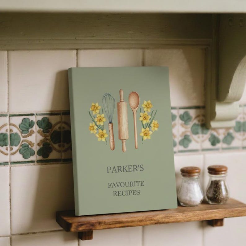 May include: A sage green recipe book with the text "PARKER'S FAVOURITE RECIPES" and illustrations of a whisk, rolling pin, and spoon, surrounded by yellow daffodils. The book is on a wooden shelf.