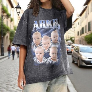 May include: Dark gray t-shirt with the word "ARKEN" in blue and white lettering. The shirt features a graphic of five babies with serious expressions, set against a blue lightning background. The shirt has a vintage, washed look.