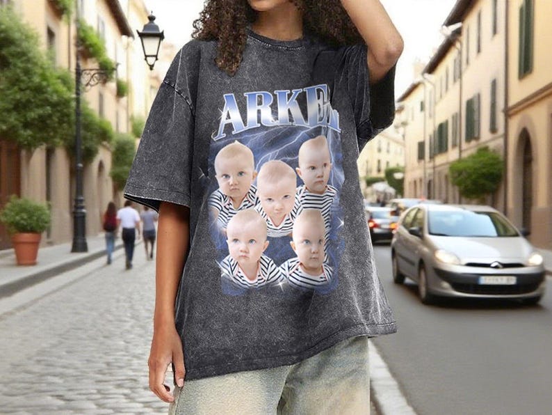 May include: Dark gray t-shirt with the word "ARKEN" in blue and white lettering. The shirt features a graphic of five babies with serious expressions, set against a blue lightning background. The shirt has a vintage, washed look.