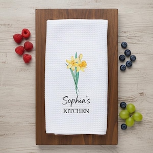 May include: White kitchen towel with a waffle weave, decorated with a watercolour illustration of yellow daffodils and the text "Sophia's KITCHEN". The towel rests on a wooden board, with raspberries, blueberries, and grapes scattered around.
