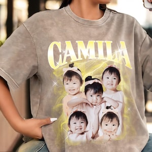 May include: A gray, short-sleeved t-shirt featuring the name "CAMILA" in a yellow, stylized font. Below the name is a graphic of five smiling children. The shirt has a faded, vintage look.