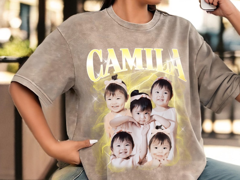 May include: A gray, short-sleeved t-shirt featuring the name "CAMILA" in a yellow, stylized font. Below the name is a graphic of five smiling children. The shirt has a faded, vintage look.