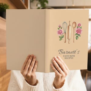 May include: A beige cookbook with the title "Barnett's Favourite Recipes" in elegant script. The cover features watercolor illustrations of a whisk, rolling pin, and spoon, framed by pink roses. The book is held open.
