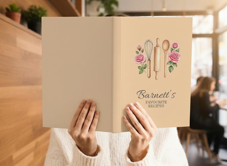 May include: A beige cookbook with the title "Barnett's Favourite Recipes" in elegant script. The cover features watercolor illustrations of a whisk, rolling pin, and spoon, framed by pink roses. The book is held open.
